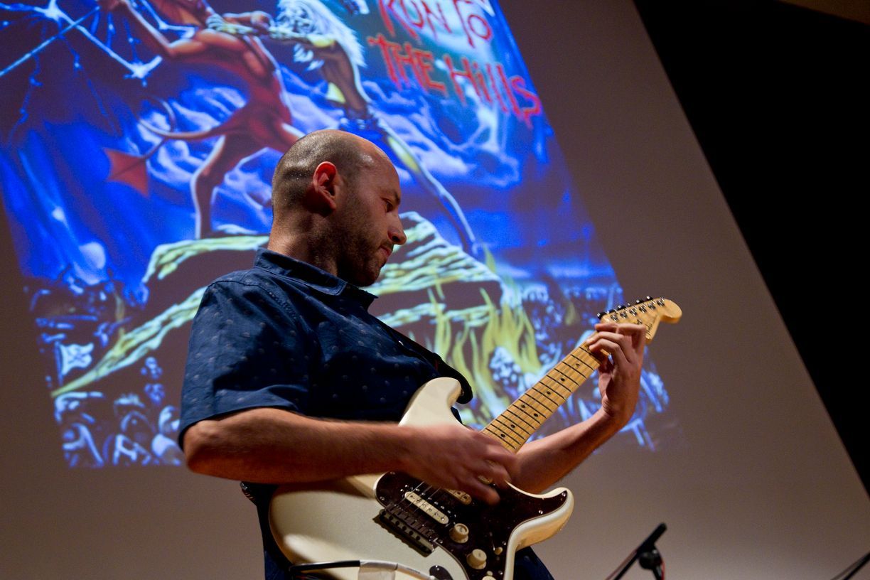 Un hombre está tocando una guitarra frente a una pantalla grande.