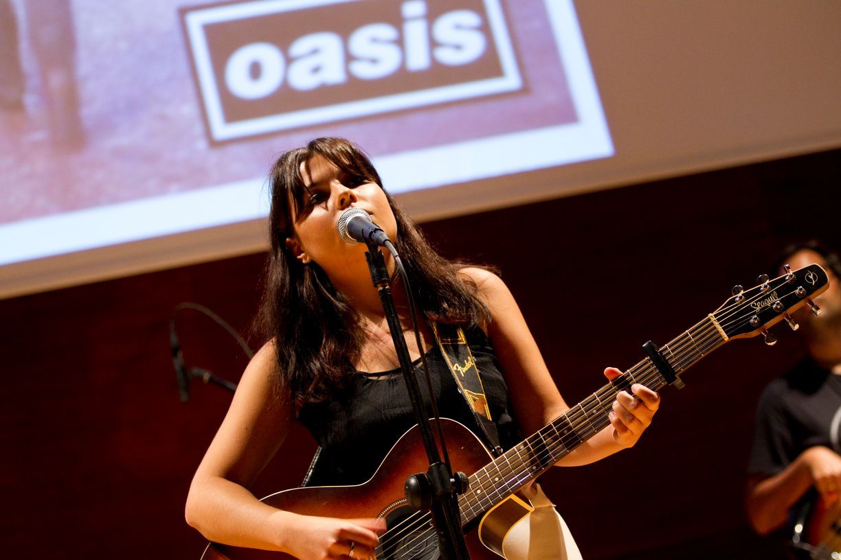 Una mujer cantando en un micrófono mientras toca una guitarra frente al logotipo de un oasis.