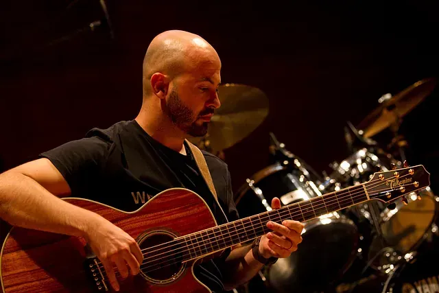 Un hombre calvo con barba está tocando una guitarra acústica en un escenario.