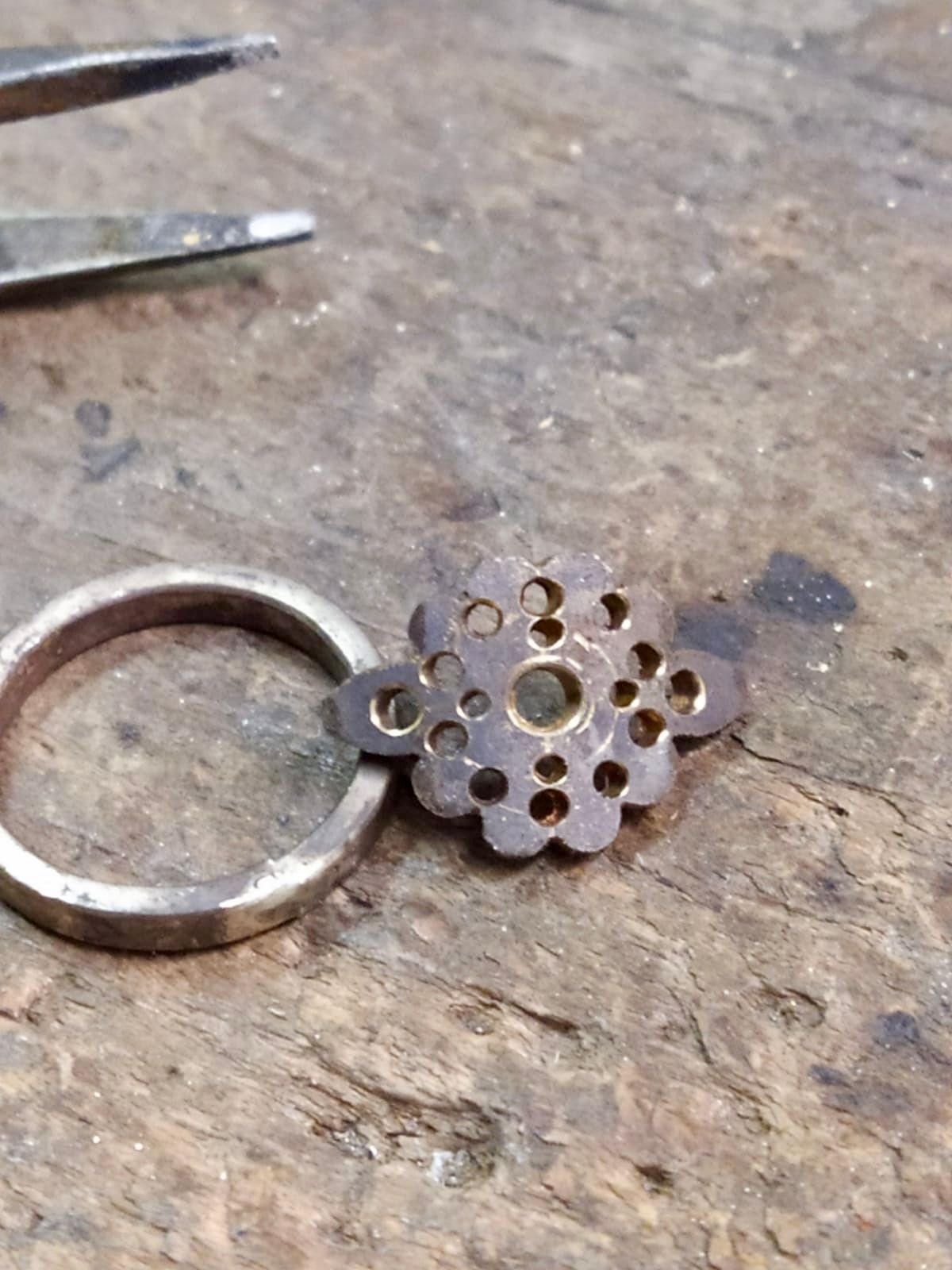 Une bague partiellement finie avec un dessus décoratif et circulaire, et une paire de pinces sur une surface de travail.