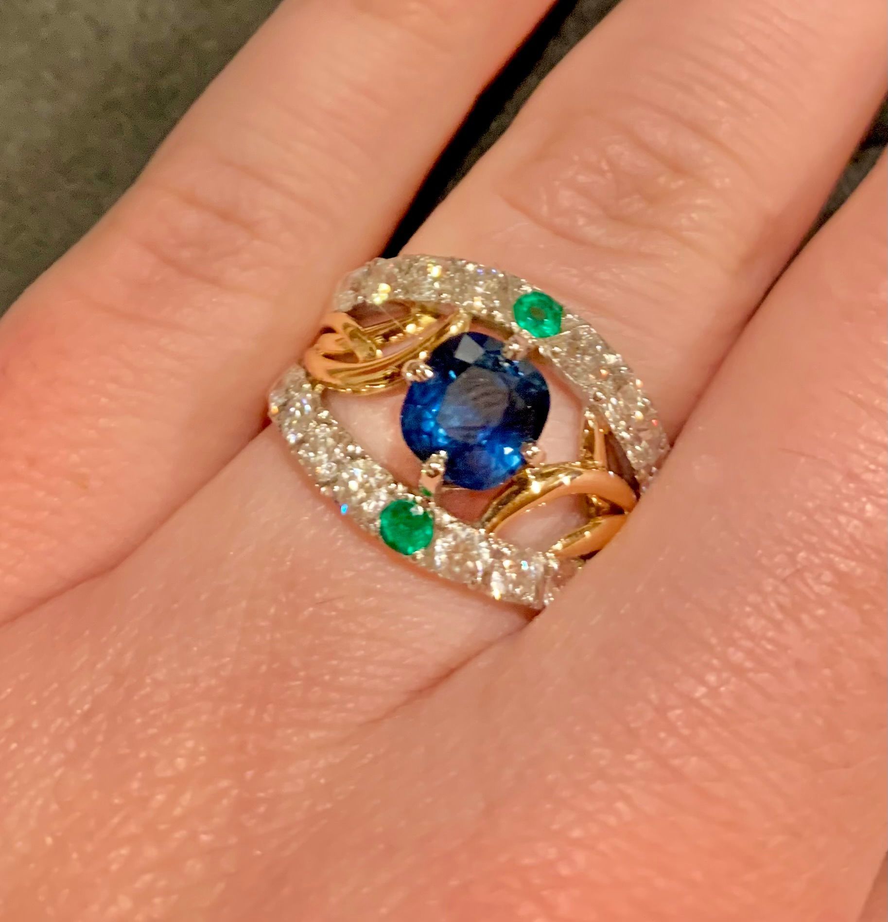 Gros plan d'une main portant une bague en or avec un gros saphir bleu, des diamants et des émeraudes.