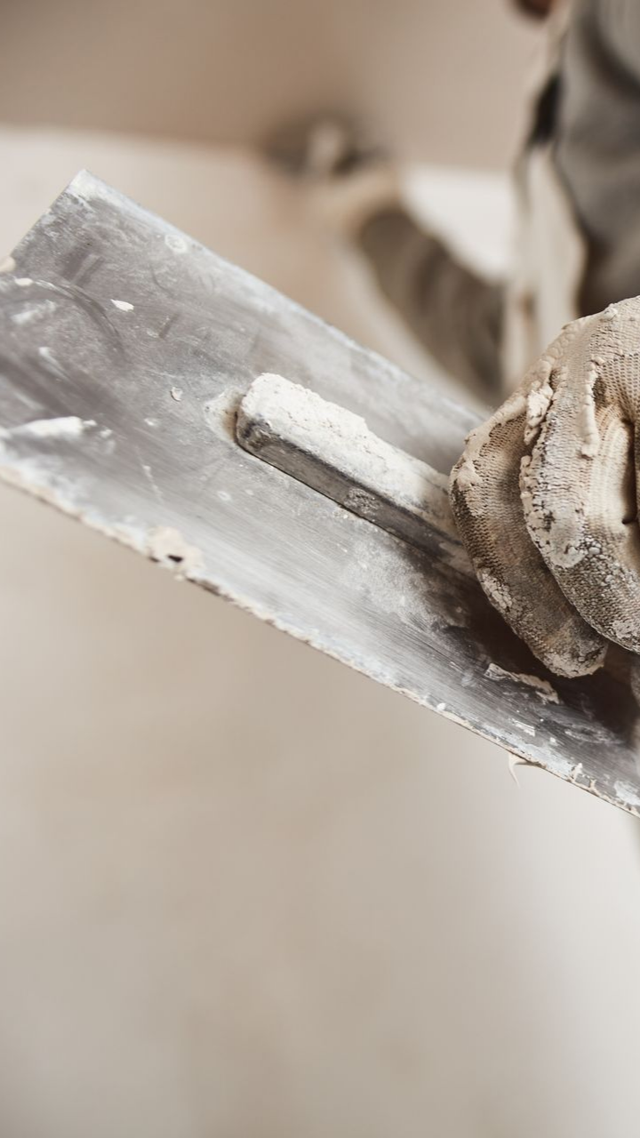 Personne tenant une truelle à la main gantée, appliquant du plâtre sur un mur.