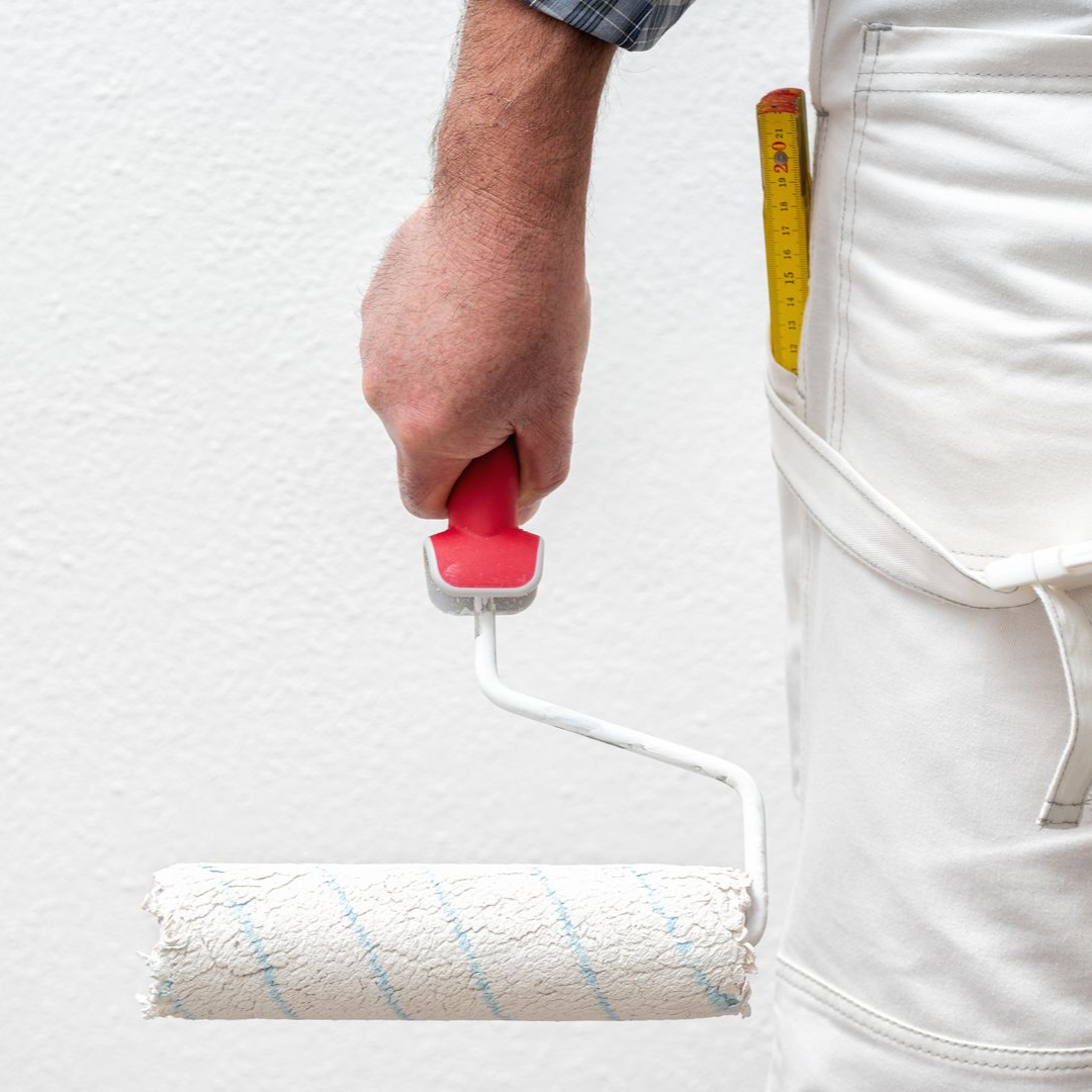 Une personne en pantalon blanc tient un rouleau à peinture contre un mur blanc.