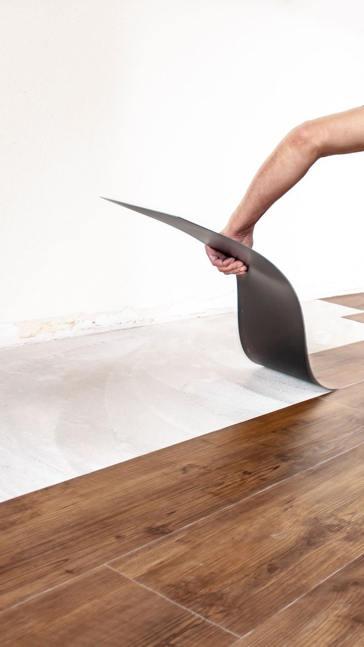 Pose du revêtement de sol : une personne soulève une bâche grise, révélant le support blanc. Des lames brunes sont posées.