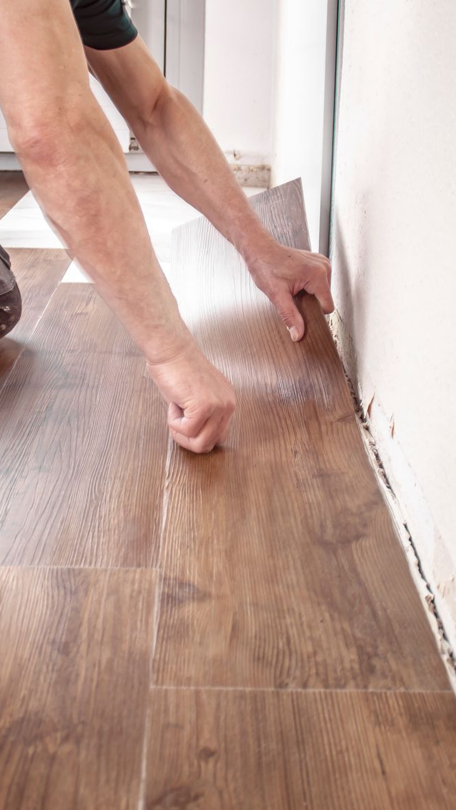 Une personne installe un revêtement de sol imitation bois à côté d'un mur blanc.