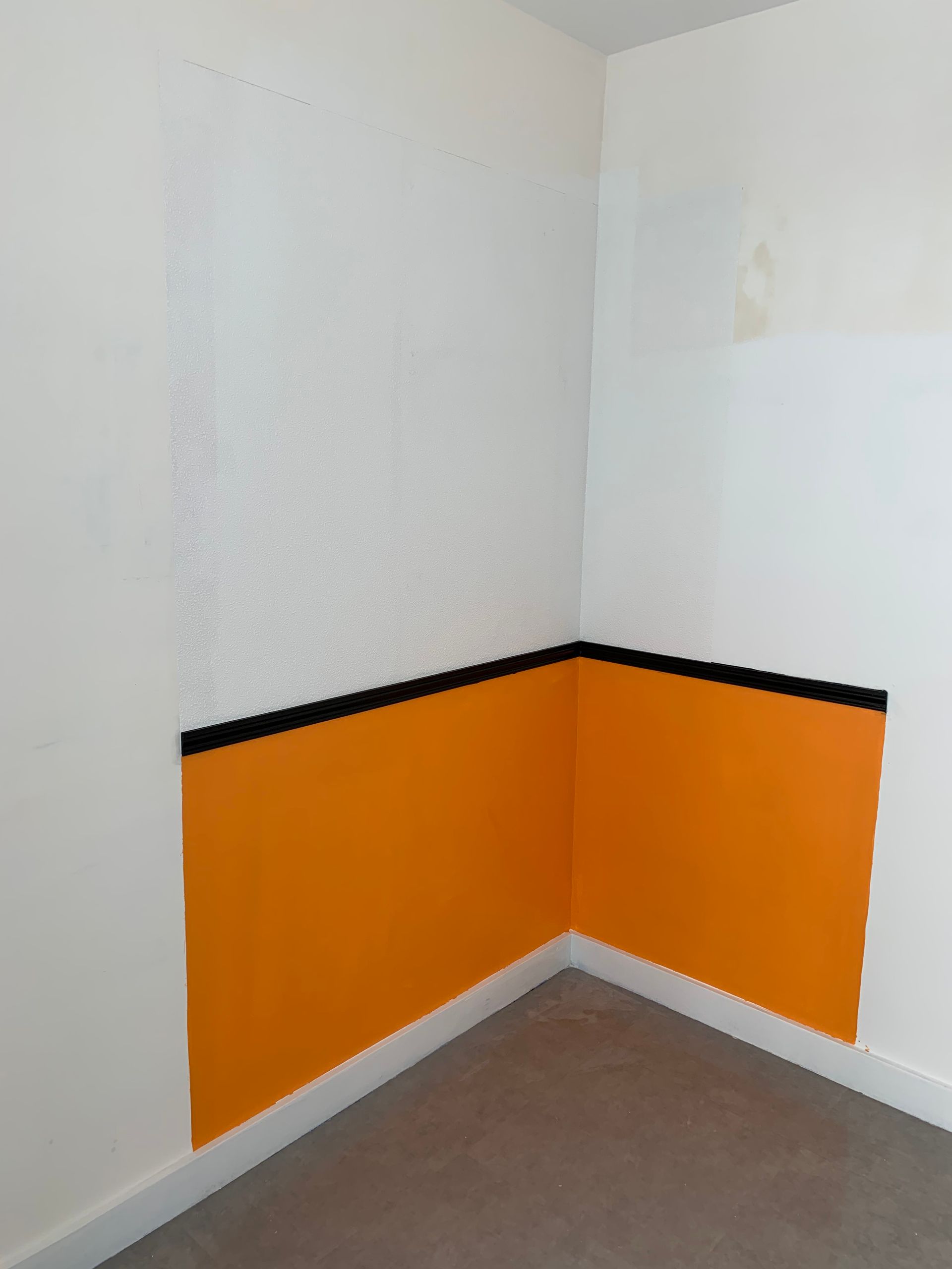 Coin d'une pièce aux murs blancs, lambris orange, moulures noires et moquette grise.