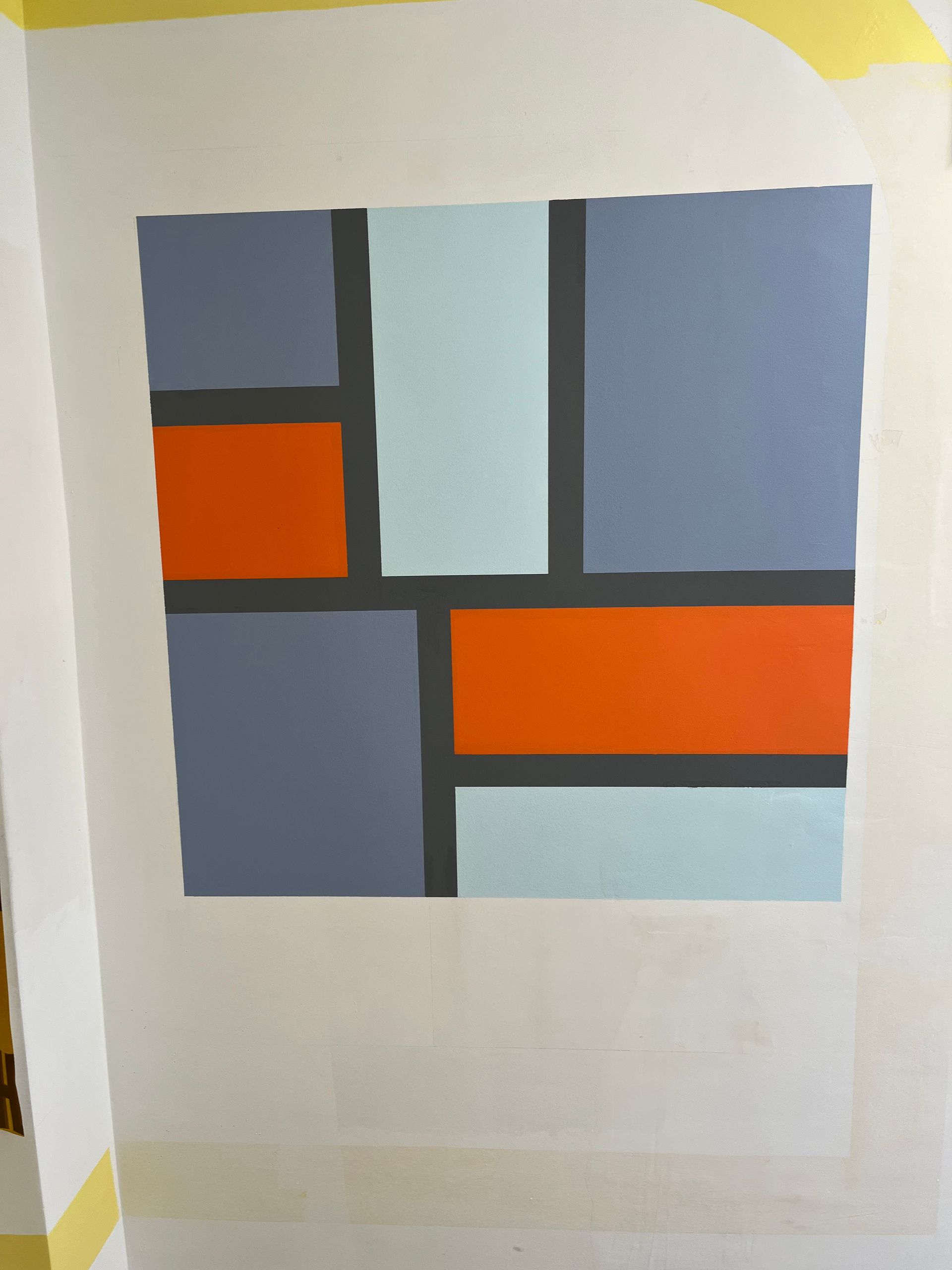 Peinture géométrique sur un mur blanc, composée de blocs gris, orange et bleu clair séparés par des lignes gris foncé.