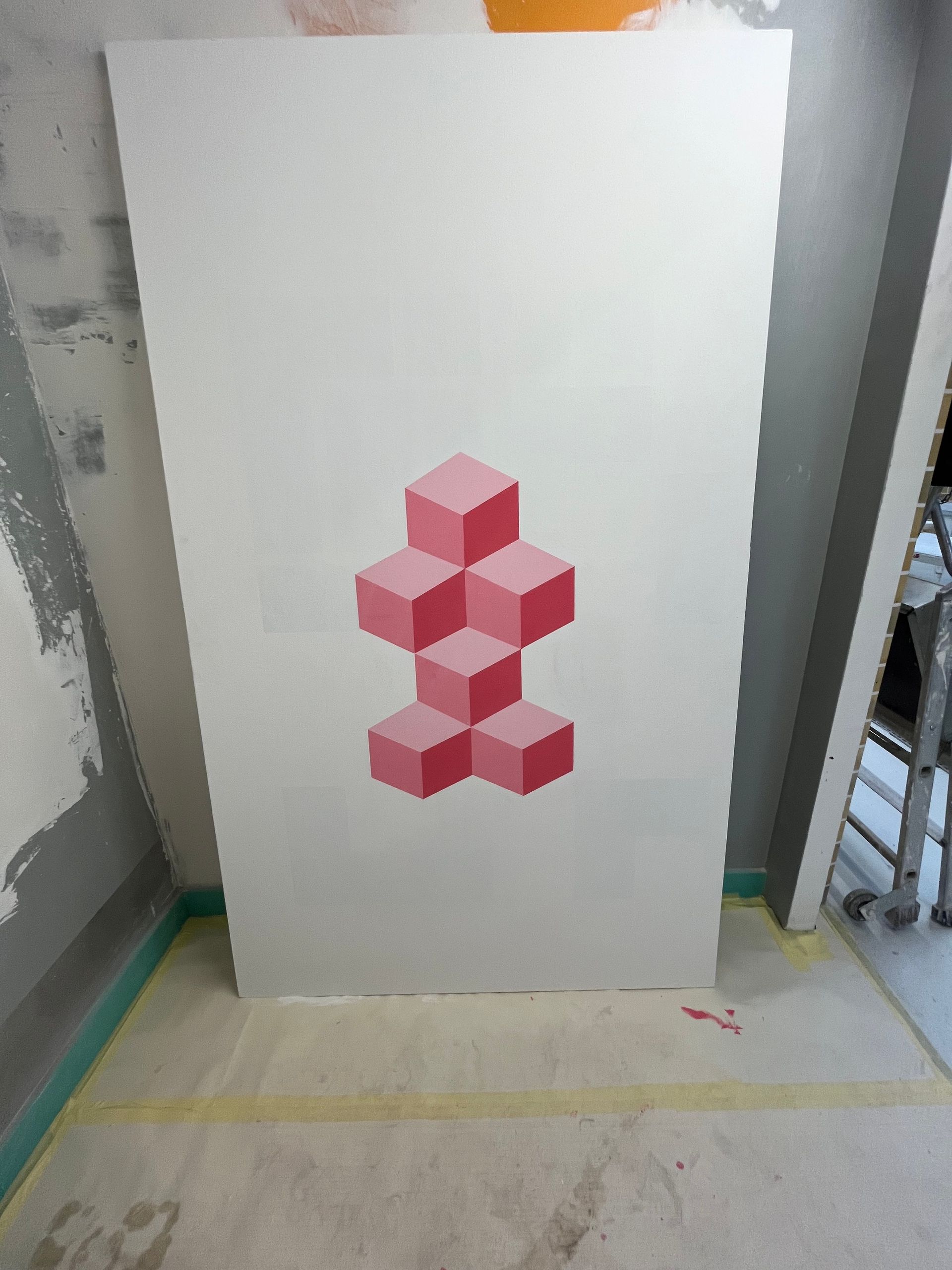 Sculpture géométrique cubique rose sur un tableau blanc appuyé contre un mur gris.