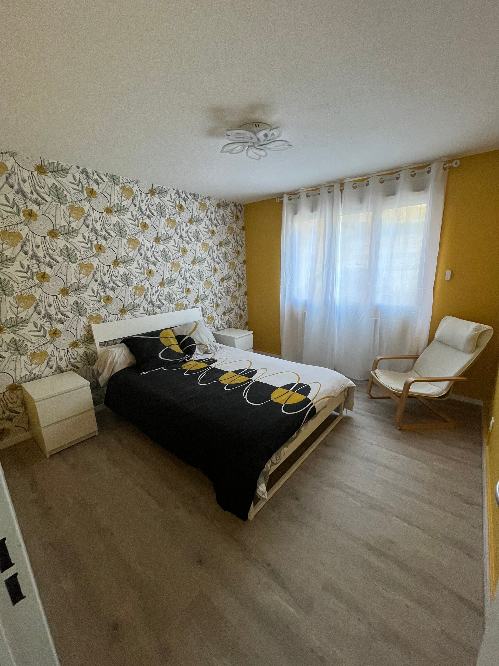 Chambre avec un lit, du papier peint à fleurs, des murs jaunes, une chaise et une fenêtre avec des rideaux.