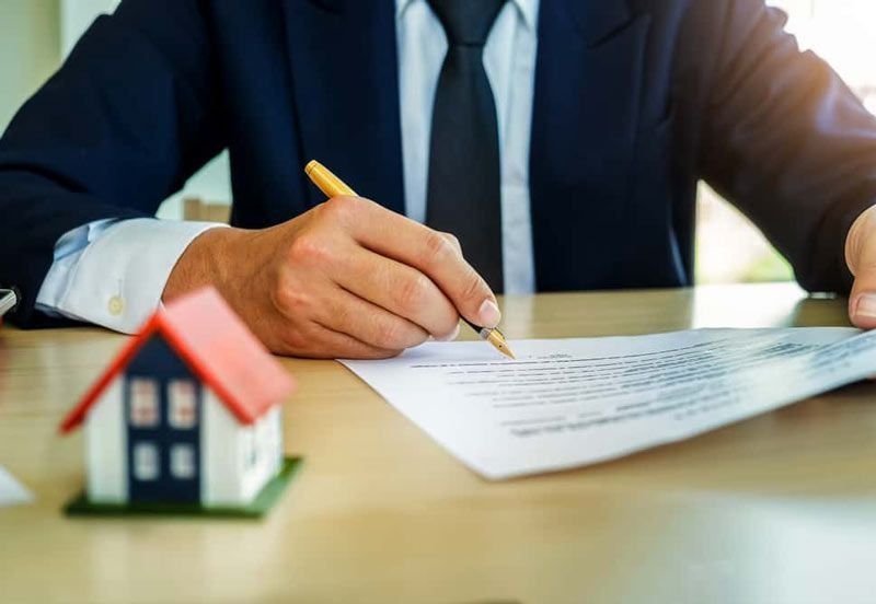 Persona de traje firmando un documento con una casa en miniatura sobre la mesa.