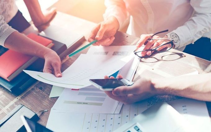 Personas colaborando en una mesa de madera, revisando documentos y usando un teléfono y una computadora portátil.