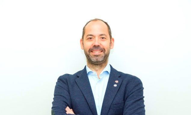 Hombre con barba, con blazer, brazos cruzados, sonriendo sobre fondo blanco.