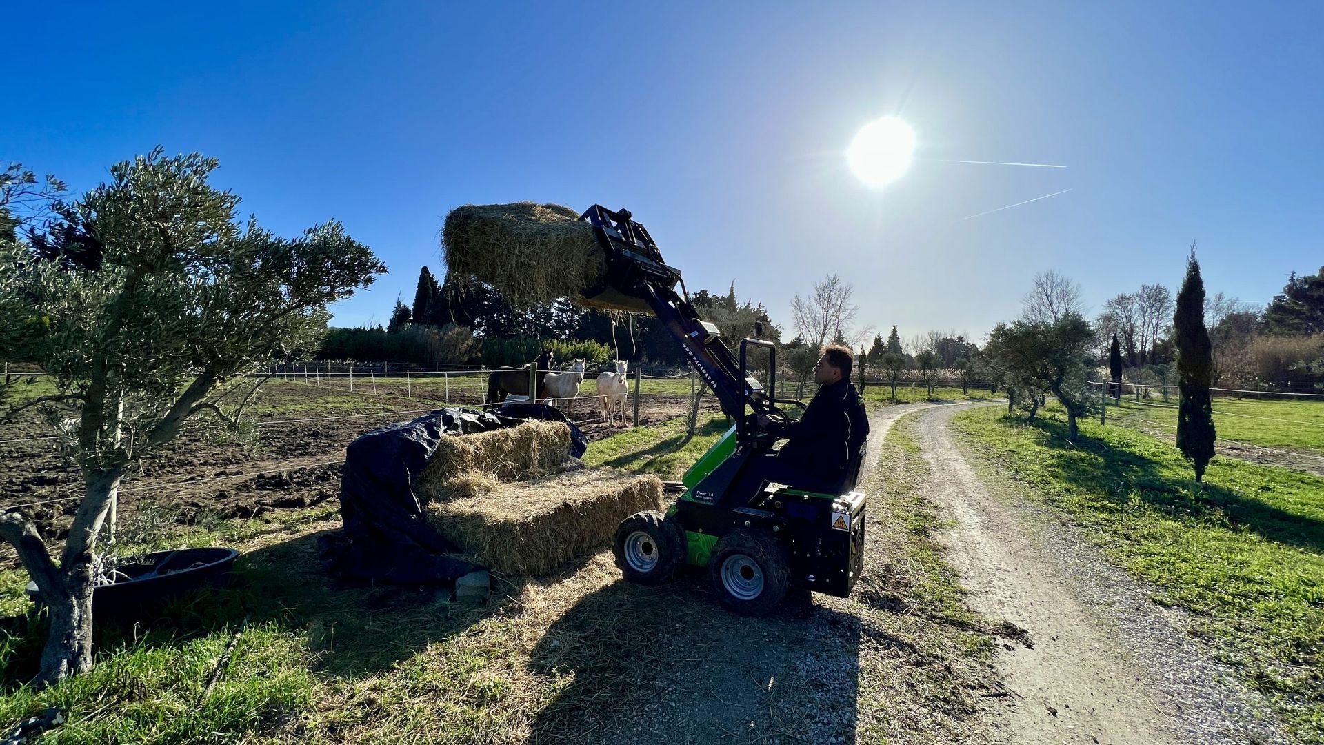 Un homme conduit un petit tracteur qui charge des balles de foin dans un champ ensoleillé.