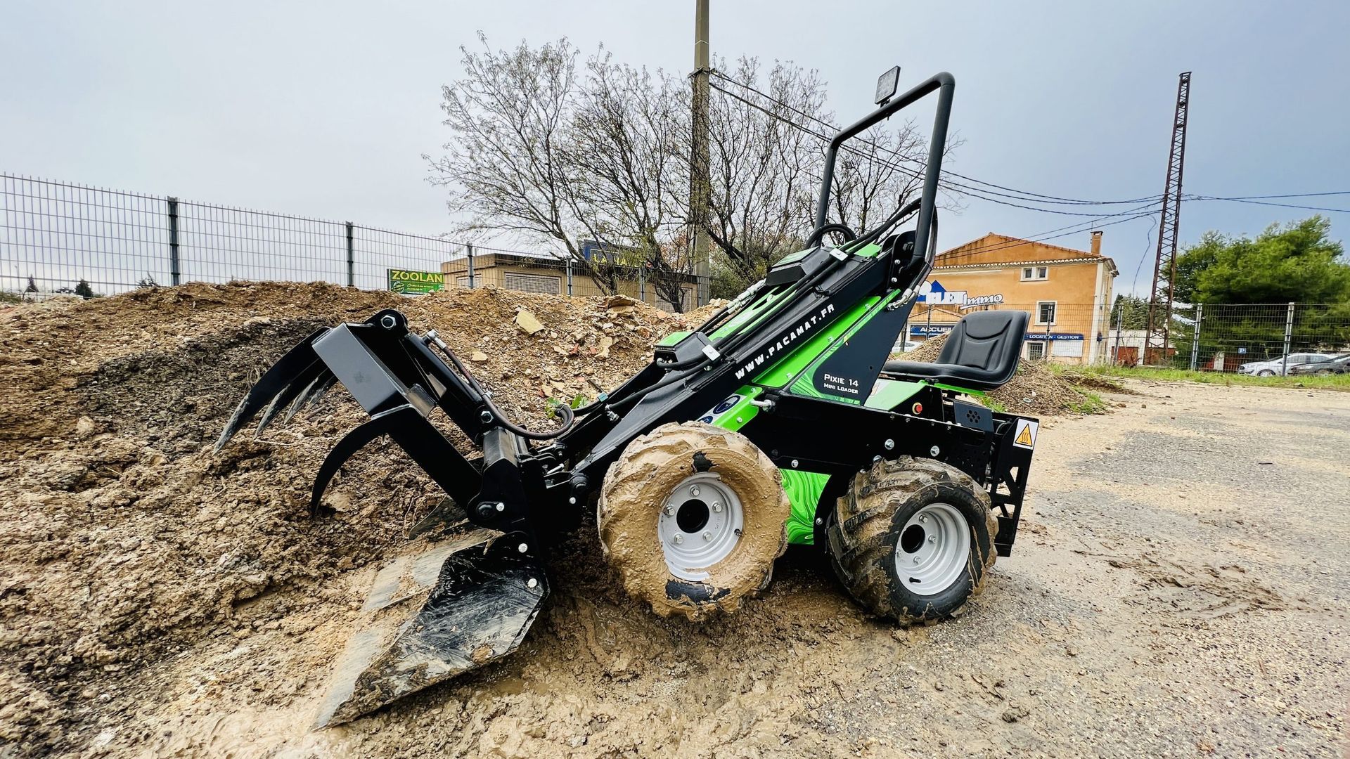 Minichargeuse verte et noire équipée d'une pince, en train de ramasser de la terre sur un chantier extérieur.