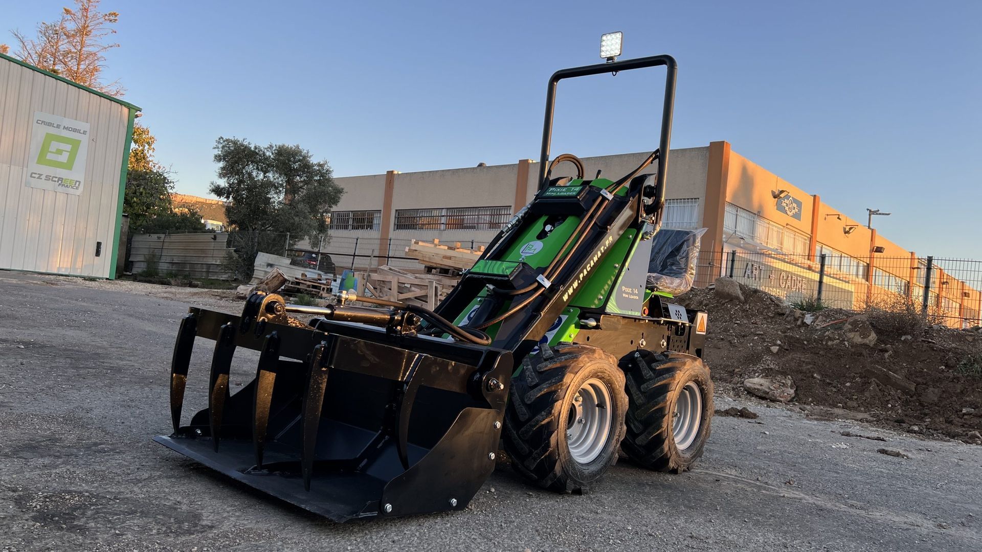 Minichargeuse verte et noire équipée d'un grappin sur du gravier devant un bâtiment.