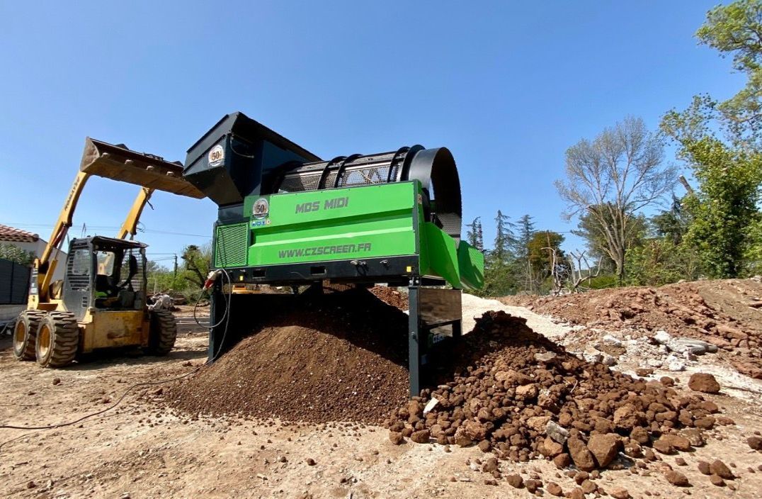 Une chargeuse compacte déverse de la terre dans un tamis à terre verte. La terre tamisée s'accumule en contrebas.