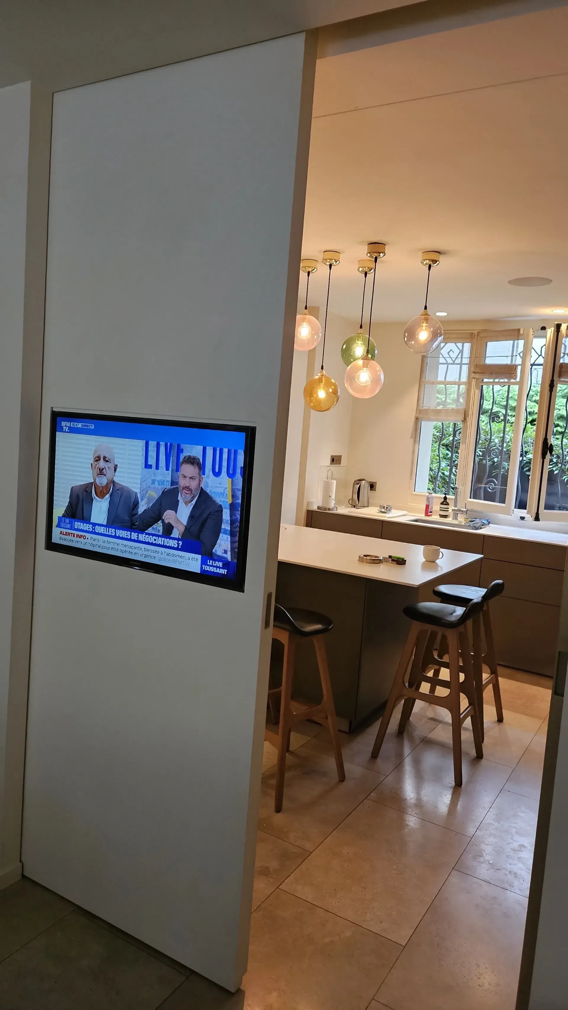 Téléviseur mural à côté d’une porte ouverte donnant sur une cuisine lumineuse avec des suspensions et des tabourets de bar