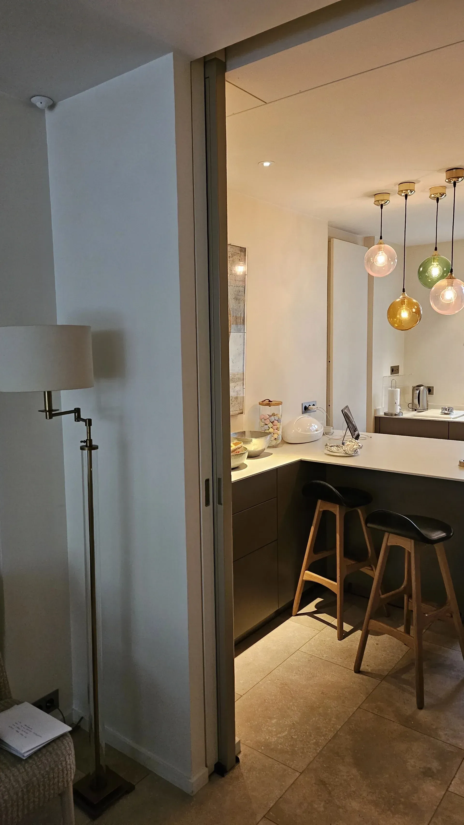 Petite cuisine d’appartement avec un plan de travail noir, des tabourets, des suspensions et une lampe blanche au premier plan