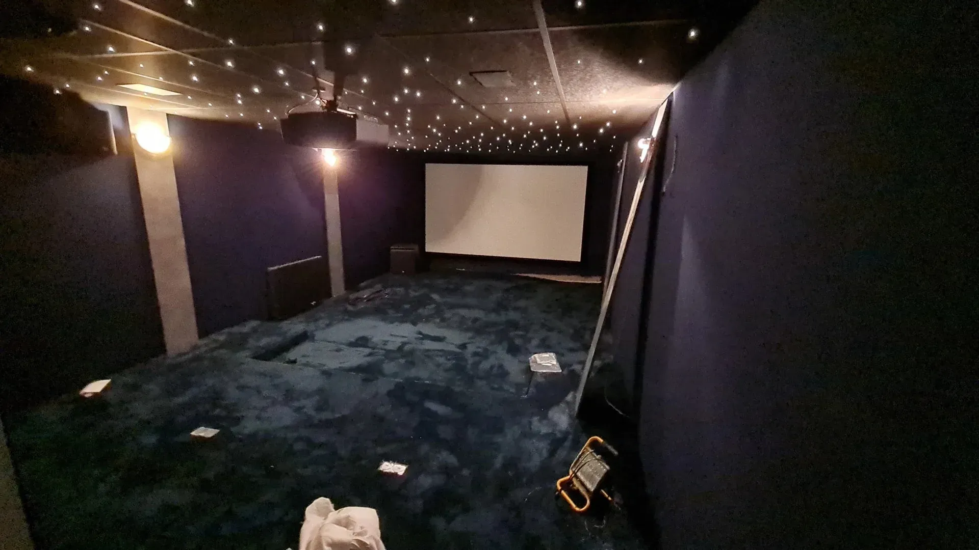 Petite salle de cinéma à domicile avec un écran de projection, un plafond illuminé de petites étoiles, une moquette foncée et le bord d’un canapé au premier plan.