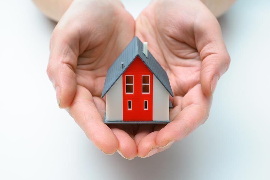 Mains jointes tenant une petite maquette de maison avec une porte rouge et un toit gris sur fond uni.