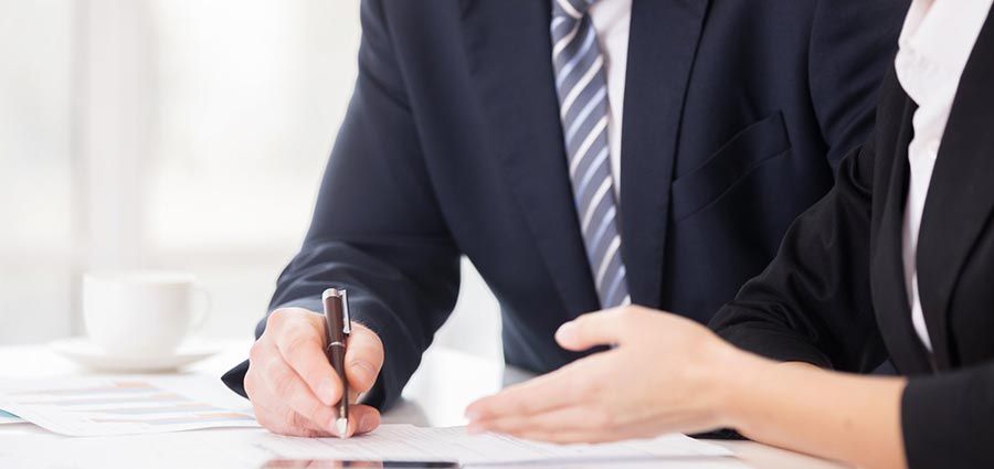 Deux hommes d'affaires en costume sont assis à un bureau ; l'un rédige un document tandis que l'autre gesticule au cours d'une réunion.