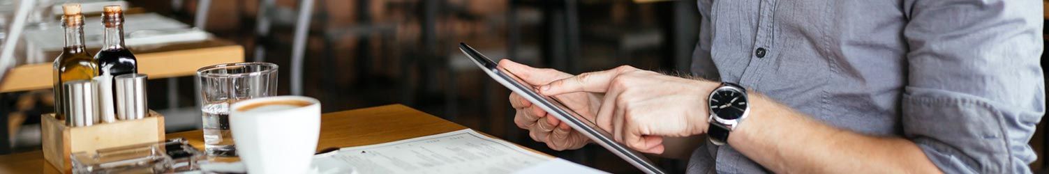 Une personne vêtue d'une chemise grise est assise à la table d'un café, une tasse de café à la main, regardant et pointant du doigt une tablette numérique.