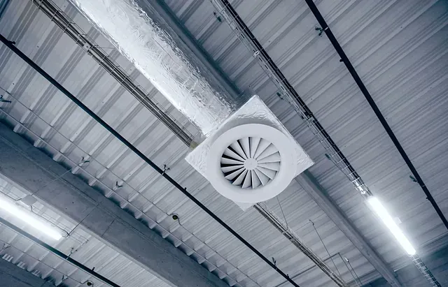 Ventilador montado en un techo de metal corrugado, probablemente en un almacén o espacio industrial. Tonos fríos y monocromáticos.