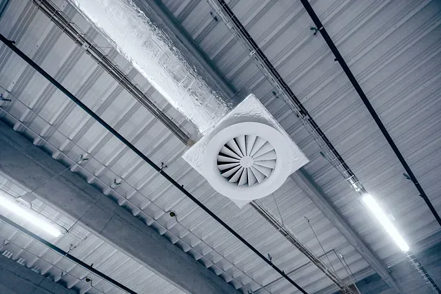 Un ventilador montado en el techo en un edificio similar a un almacén, con techo de metal e iluminación superior.
