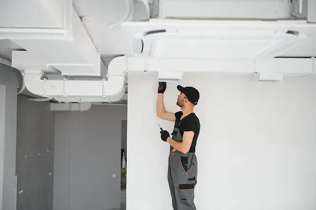 Un técnico con un mono gris instala conductos en una habitación de paredes blancas, sosteniendo un destornillador.