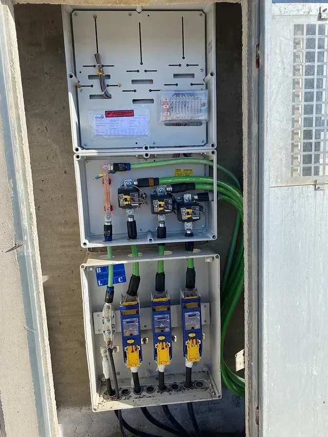 Caja eléctrica abierta con cables verdes, fusibles y medidores montados sobre una superficie de concreto.