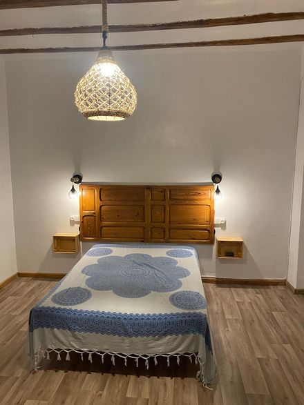 Dormitorio con cabecero de madera y mesitas de noche flotantes, cama cubierta con una manta estampada en azul y blanco y lámpara de tejido.