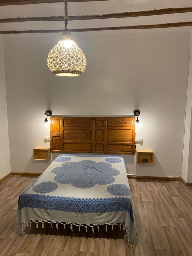 Dormitorio con cabecero de madera y mesitas de noche flotantes, cama cubierta con una manta estampada en azul y blanco y lámpara de tejido.
