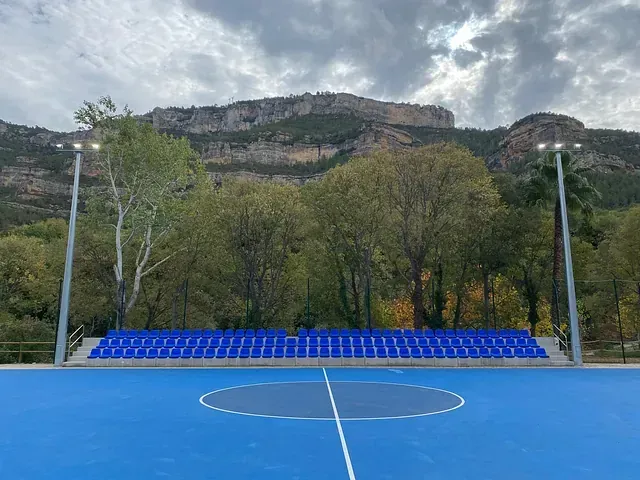 Una cancha deportiva azul con asientos azules frente a árboles y una gran montaña bajo un cielo nublado. Dos luces altas se alzan a cada lado.