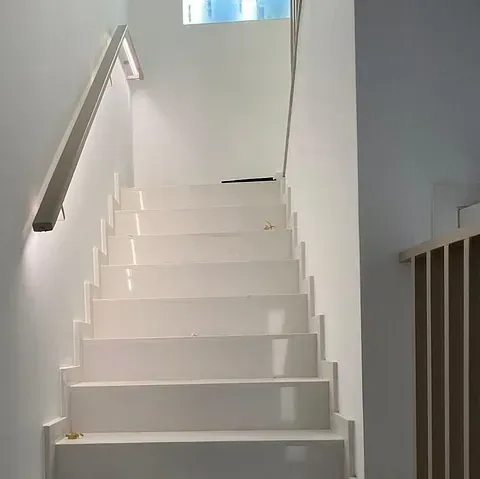Escalera blanca de diseño moderno, iluminada por una lámpara de pared. La parte superior da a una pequeña ventana.