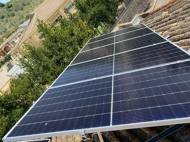 Paneles solares instalados en un tejado de tejas, probablemente para generar electricidad. Al fondo se ve vegetación y un edificio.