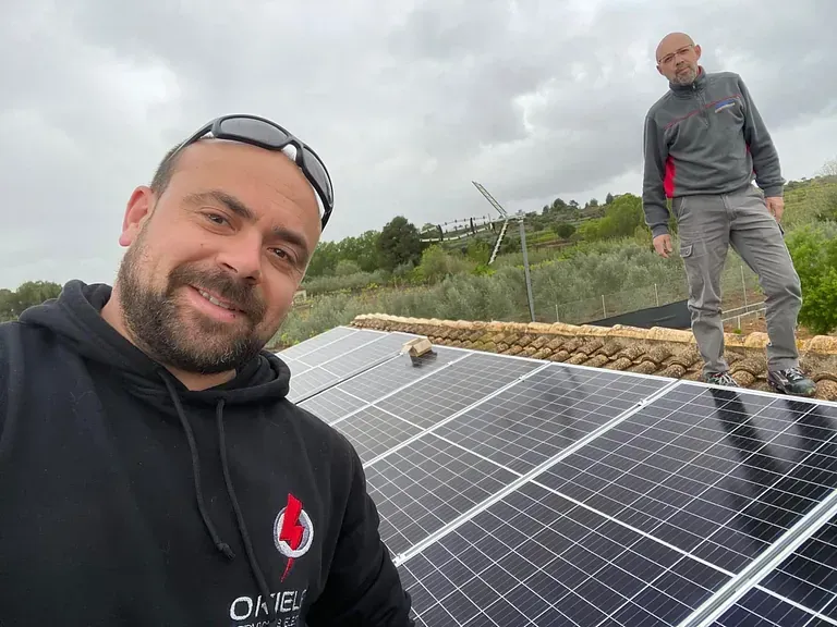 Dos hombres instalando paneles solares en un tejado. Uno se toma una selfi; el otro está cerca. Día nublado con un fondo rural.