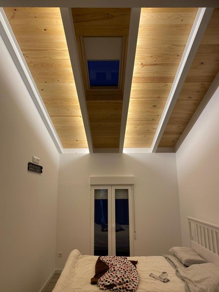 Interior de dormitorio con techo de vigas de madera, claraboya y cama con sábanas arrugadas. Paredes blancas e iluminación tenue.