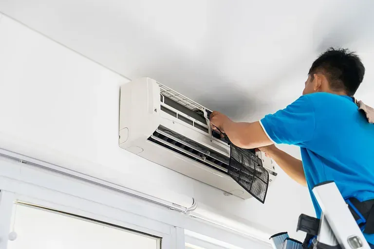 Hombre con camisa azul limpiando un filtro de aire acondicionado, de pie en una escalera cerca de una ventana en una habitación blanca.