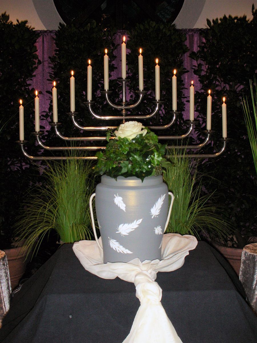 Eine graue Urne mit Federn darauf steht vor einer Menora