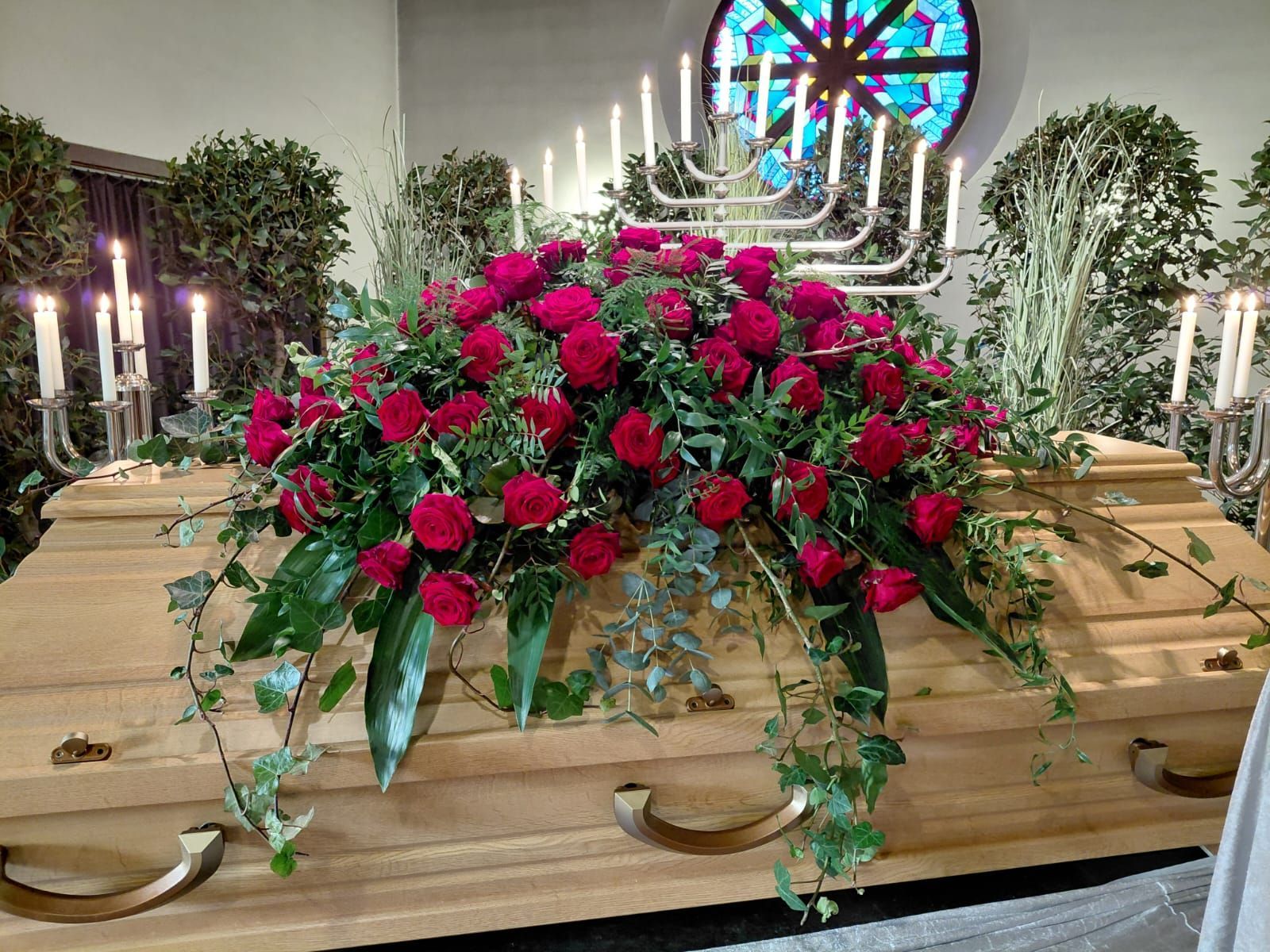 Blumenschmuck aus roten Rosen auf einem Holzsarg, umgeben von brennenden Kerzen in einer Kirche