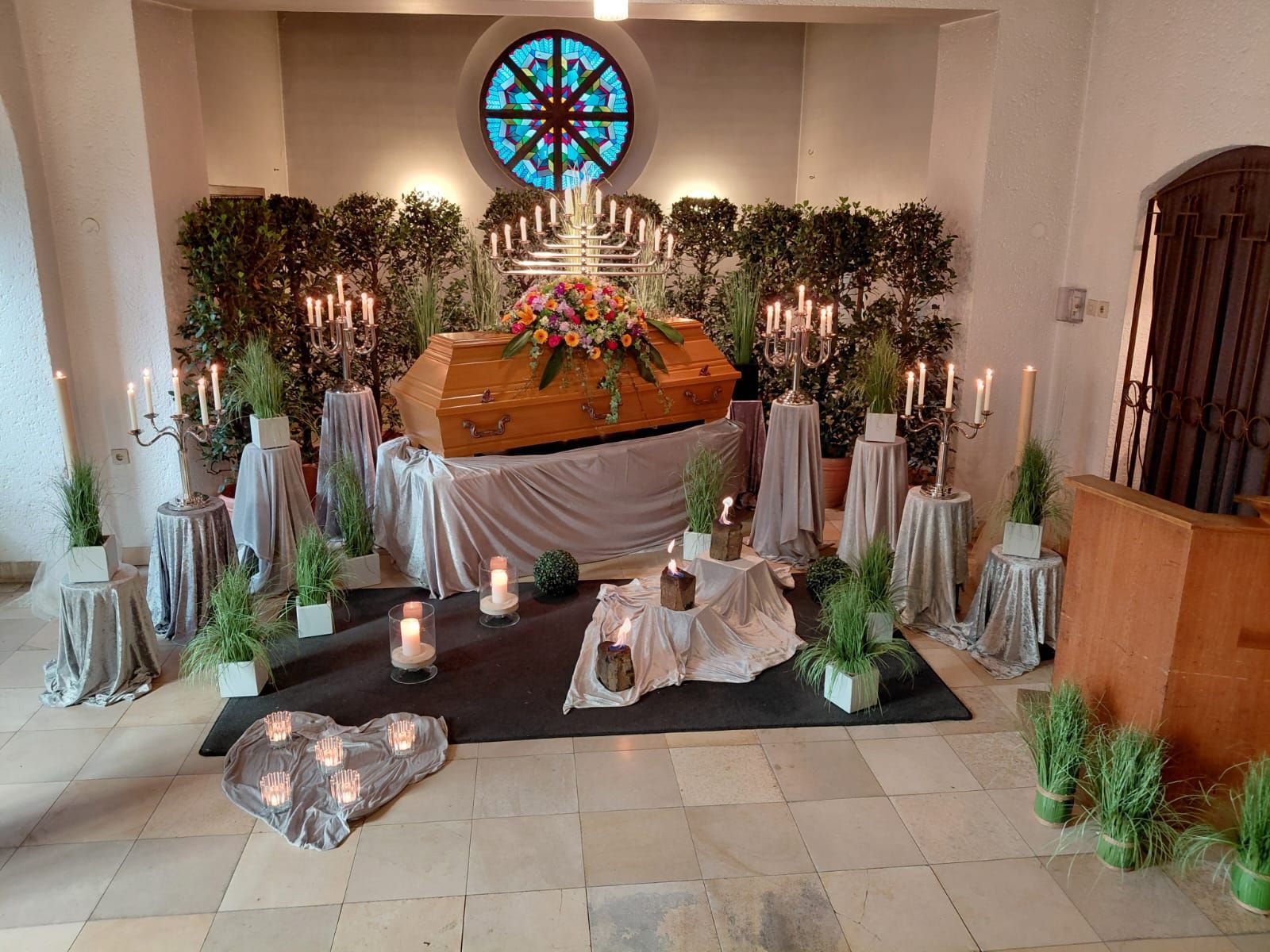 Geschmückter Hausaltar mit Blumen, Kerzen und Pflanzen, darüber ein Buntglasfenster