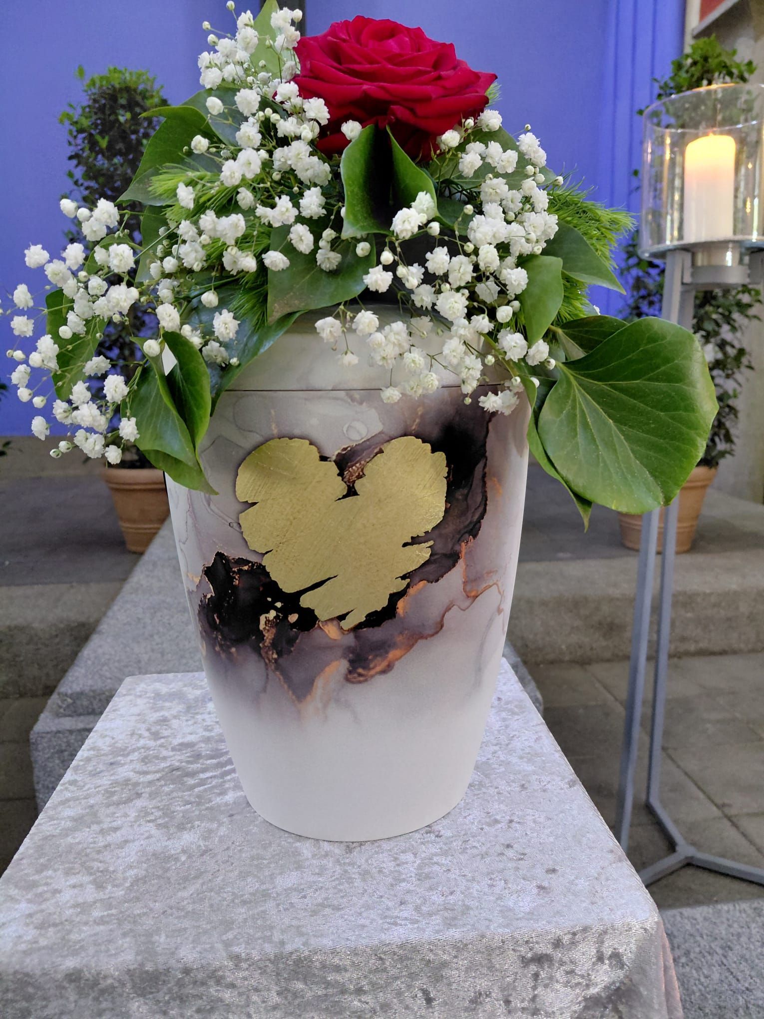 Weiße Gedenkkerze mit Rosen, Schleierkraut und einem goldenen Herzen auf einem Steinsockel