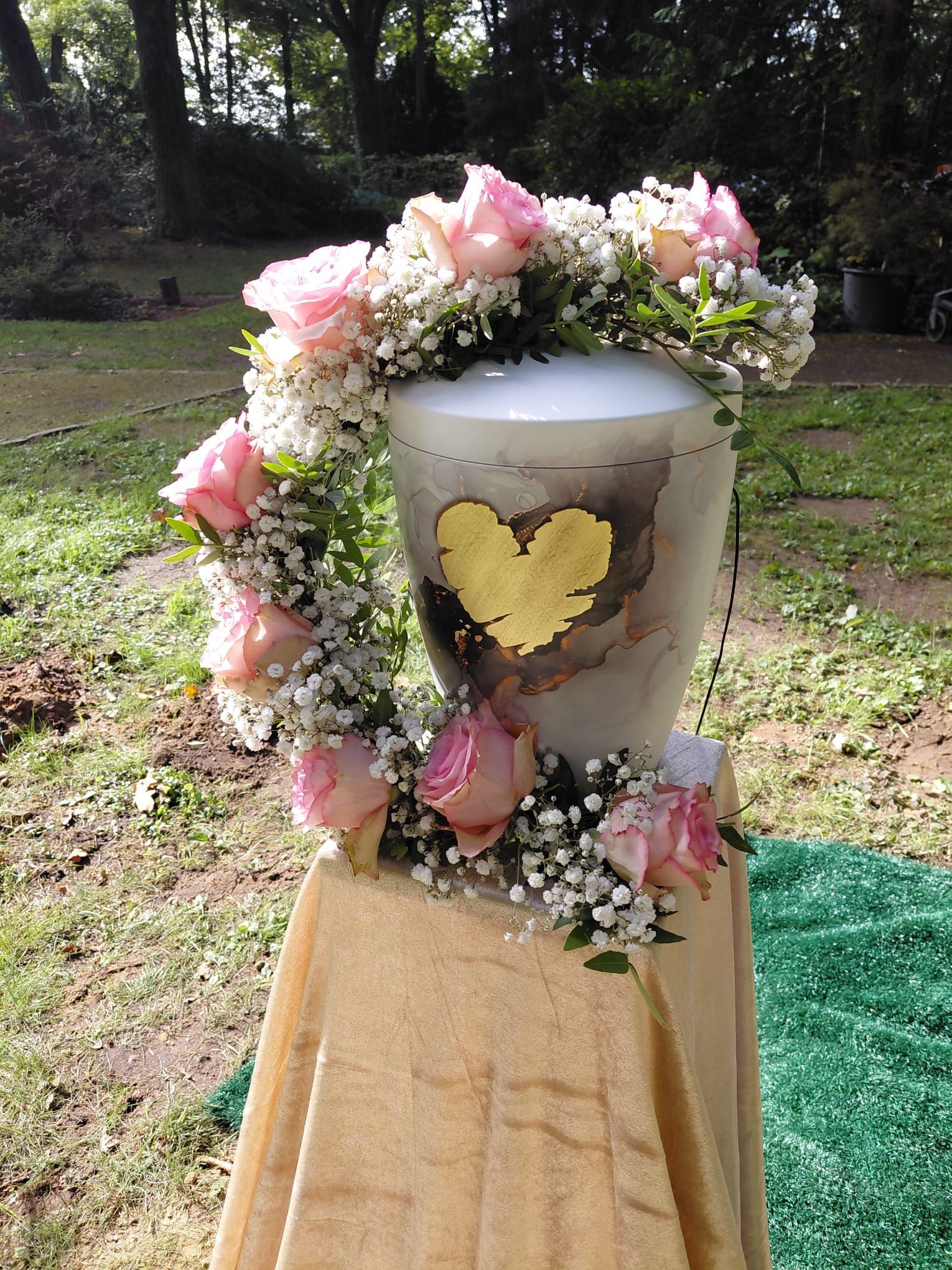Weiße Urne mit rosa und weißen Blumen und einem goldenen Herz, im Freien auf einem Holzständer