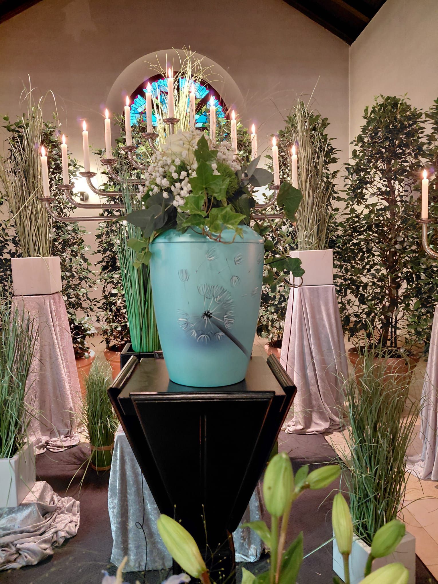 Blauer Pflanzkübel mit weißen Blumen auf einem Kirchenaltar, flankiert von Grünpflanzen und Kerzen in einem Kirchenraum mit Buntglasfenstern