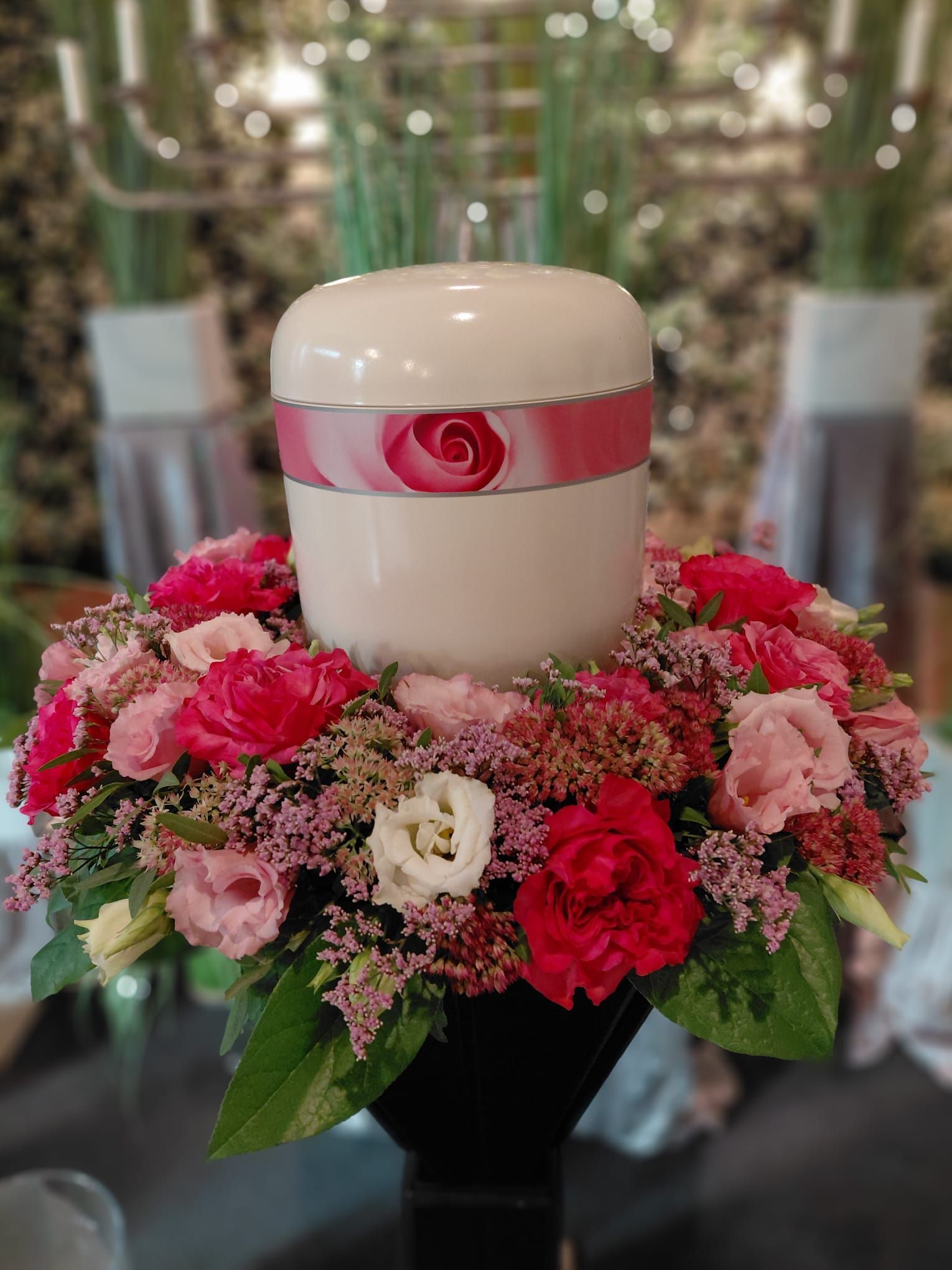 Weiße Urne mit rosa Schleife auf einem rot-rosa Blumengesteck