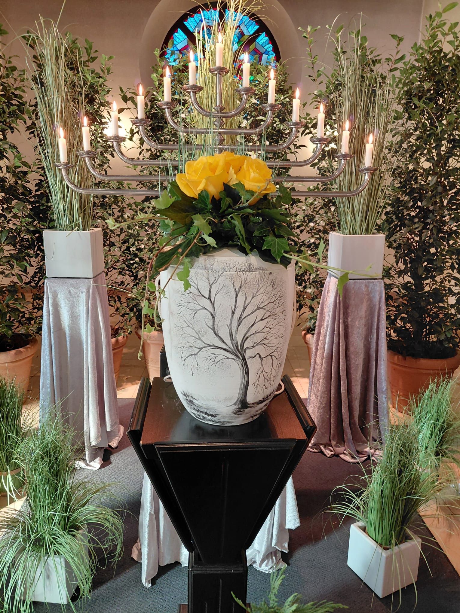 Blumenarrangement mit gelben Blumen in einer großen weißen Vase, umgeben von Grün und Kerzen in einer Kirche