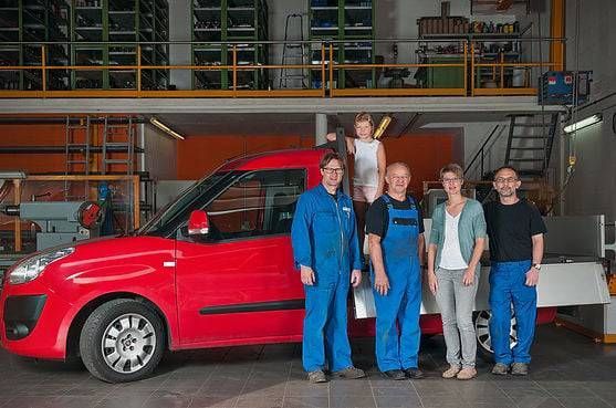 Fünf Personen stehen in einer Werkstatt neben einem roten Auto. Ein Kind sitzt auf dem Autodach, die anderen lächeln.