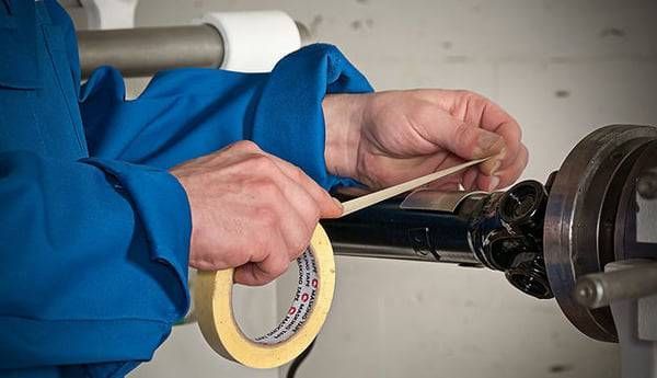 Eine Person in einem blauen Overall klebt einen schwarzen zylindrischen Gegenstand mit Klebeband ab.