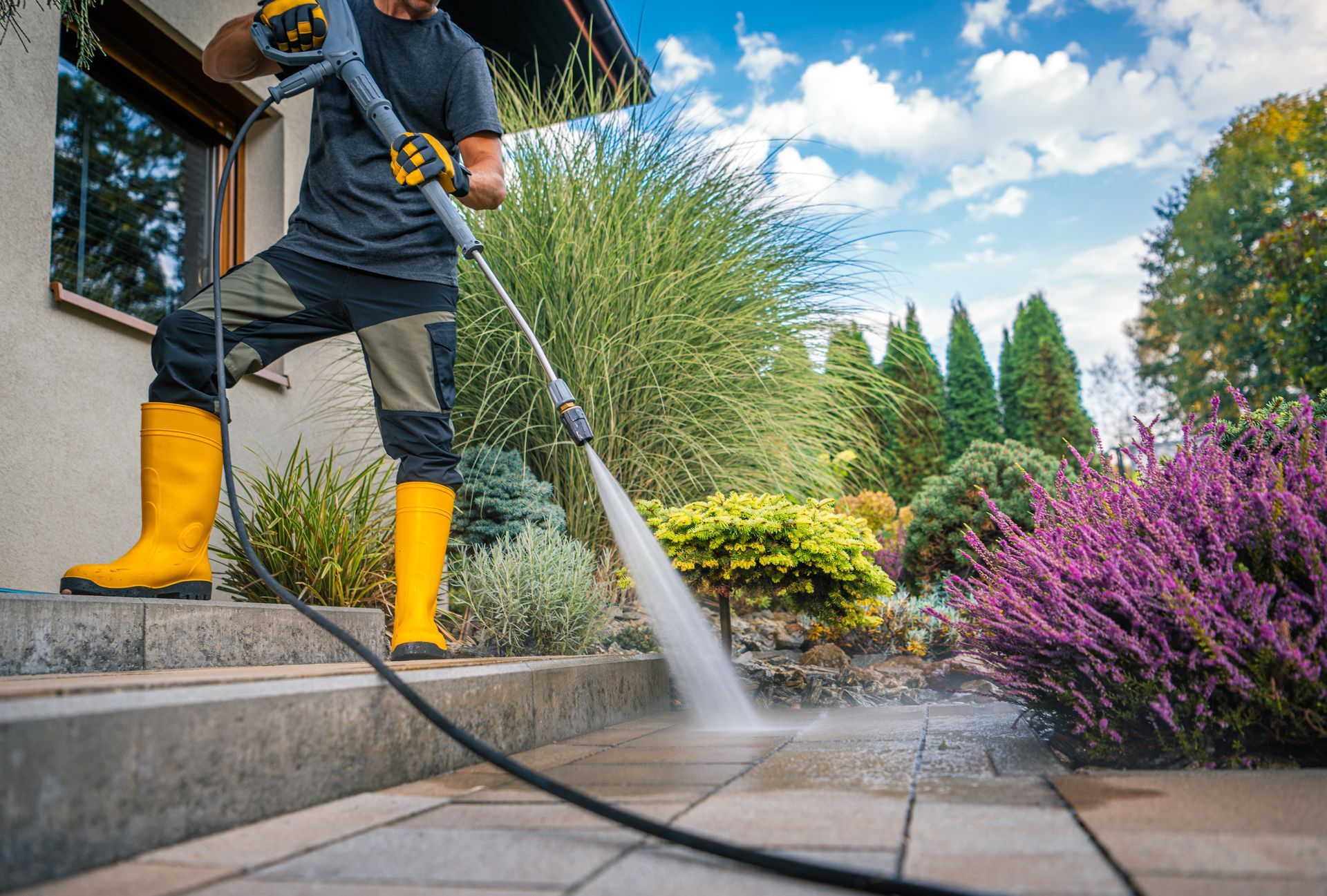Personne nettoyant un patio avec un nettoyeur à haute pression, bottes jaunes, plantes, ciel bleu.