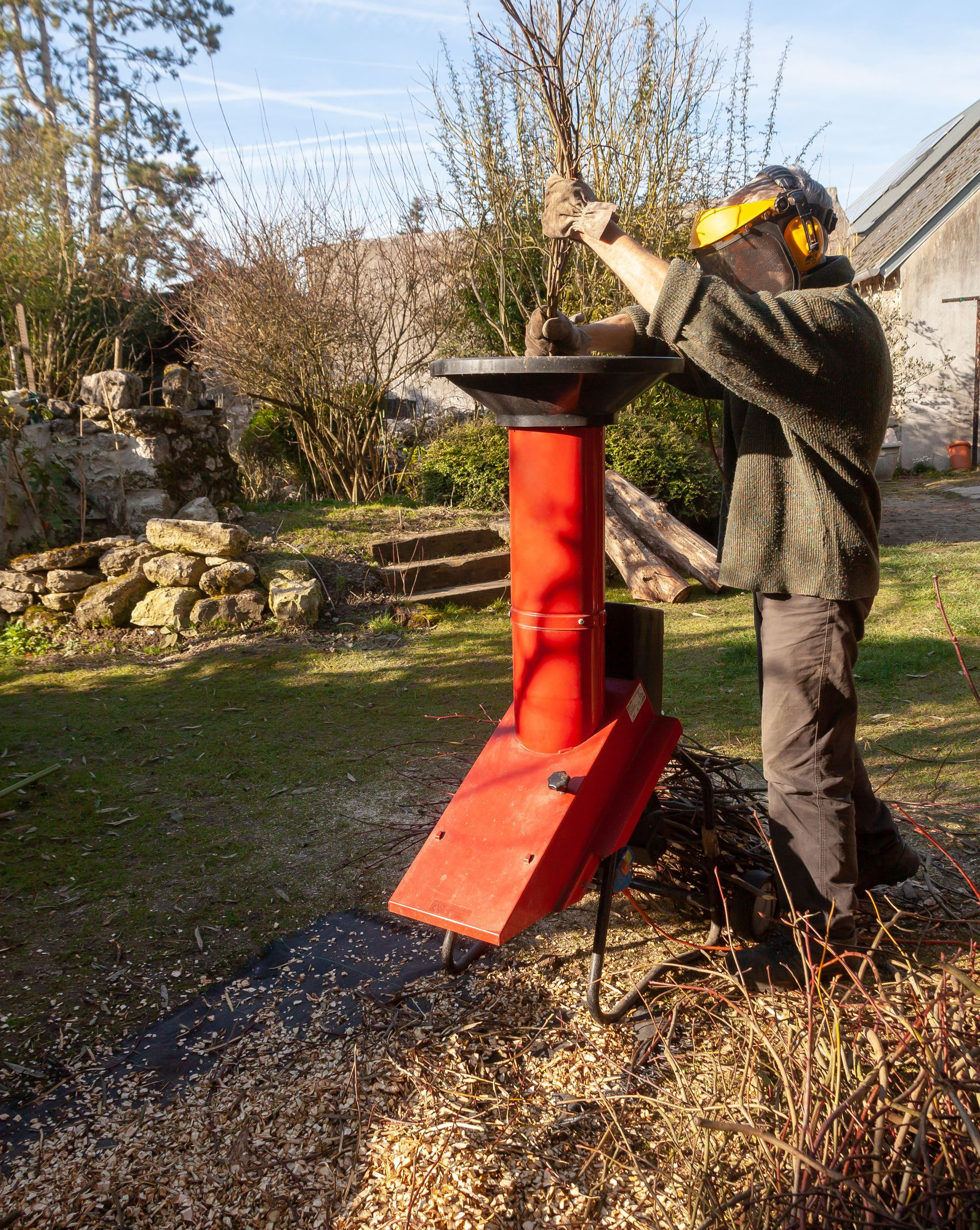 Un homme alimente un broyeur à bois rouge avec des branches dans une cour ensoleillée.